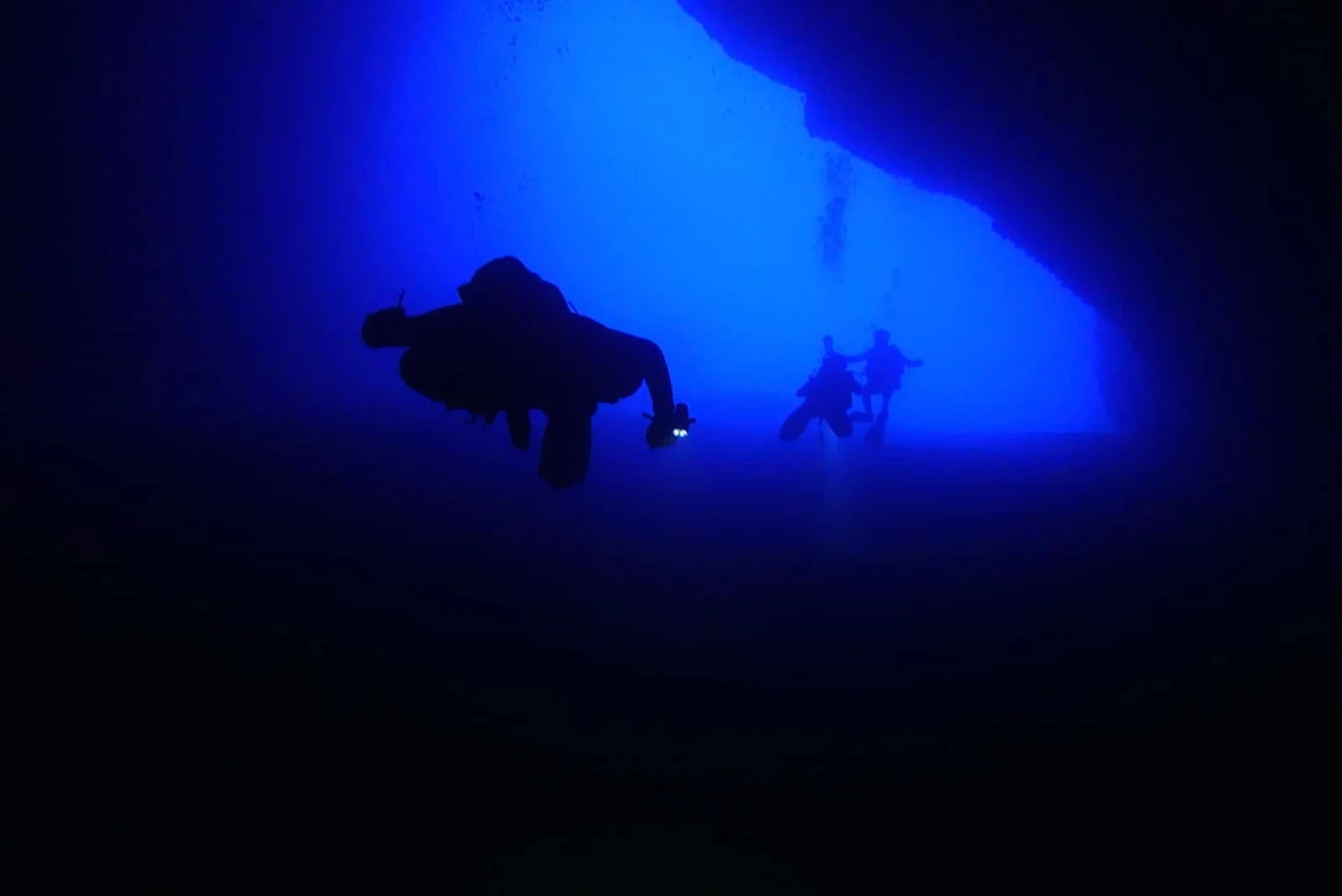 Taucher im Blauwasser vor einer Wand, aus dem Dunkeln heraus fotografiert